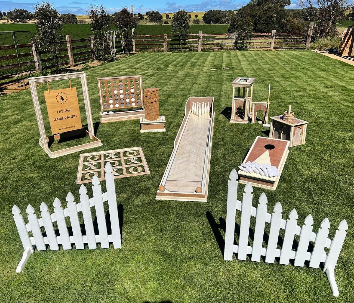 Wooden outdoor games like cornhole and tic-tac-toe arranged on a green lawn with white fences.