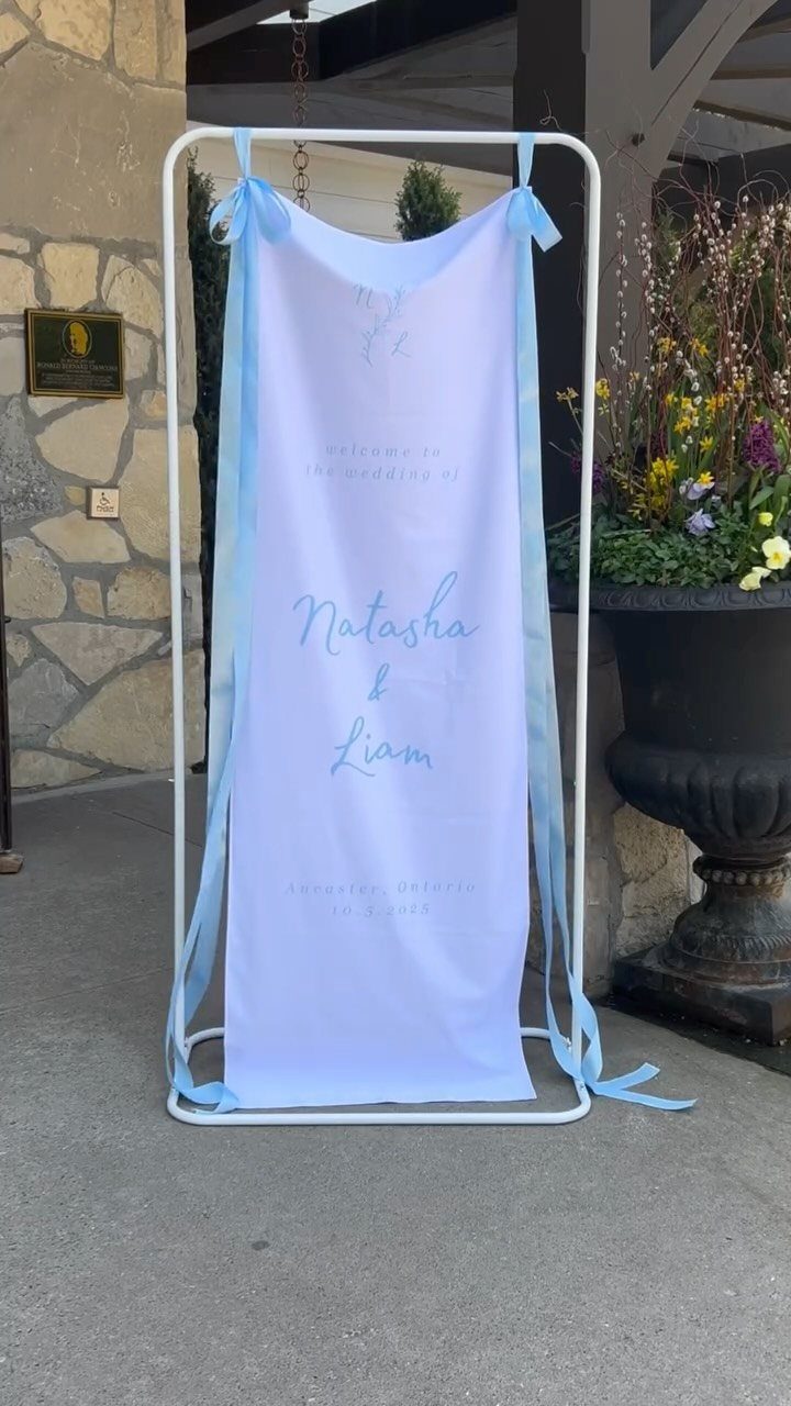 A light blue fabric wedding welcome banner hanging on a white metal stand outdoors.