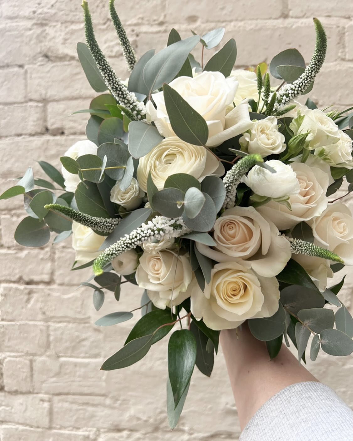 A bridal bouquet with white roses, cream ranunculus, and eucalyptus leaves held against a brick wall.