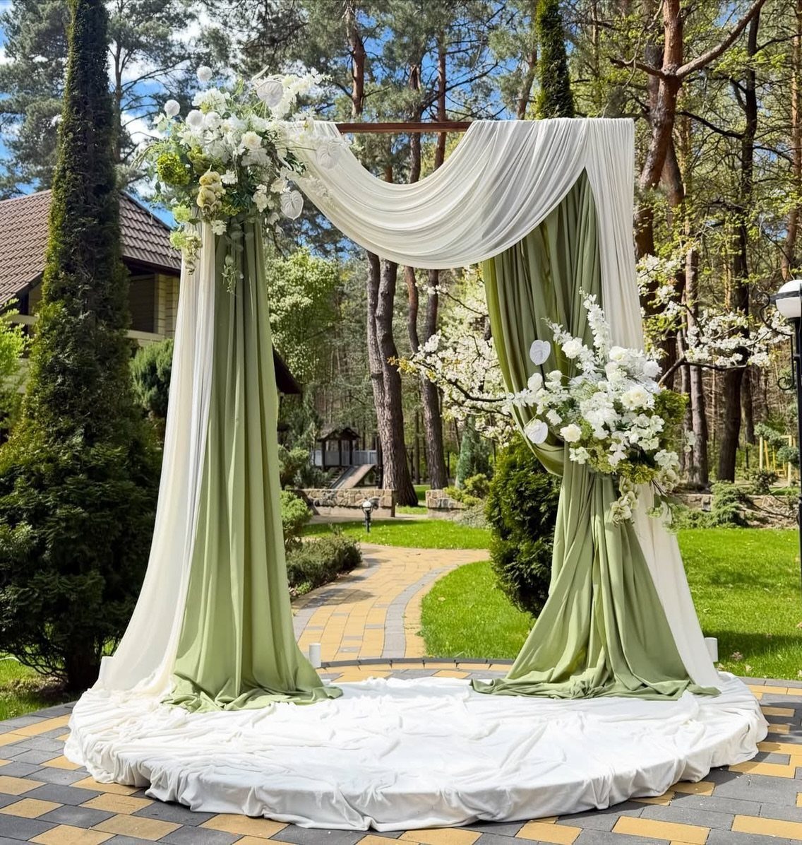 A wedding ceremony arch with sage green and white fabric draped with white floral arrangements.