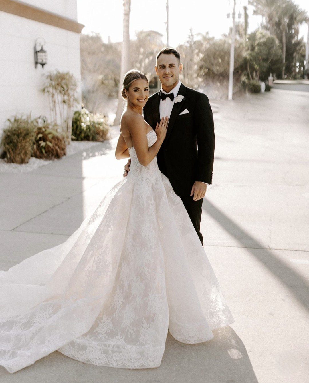 A bride wearing a strapless lace wedding dress and a groom in a tuxedo posing together outdoors.