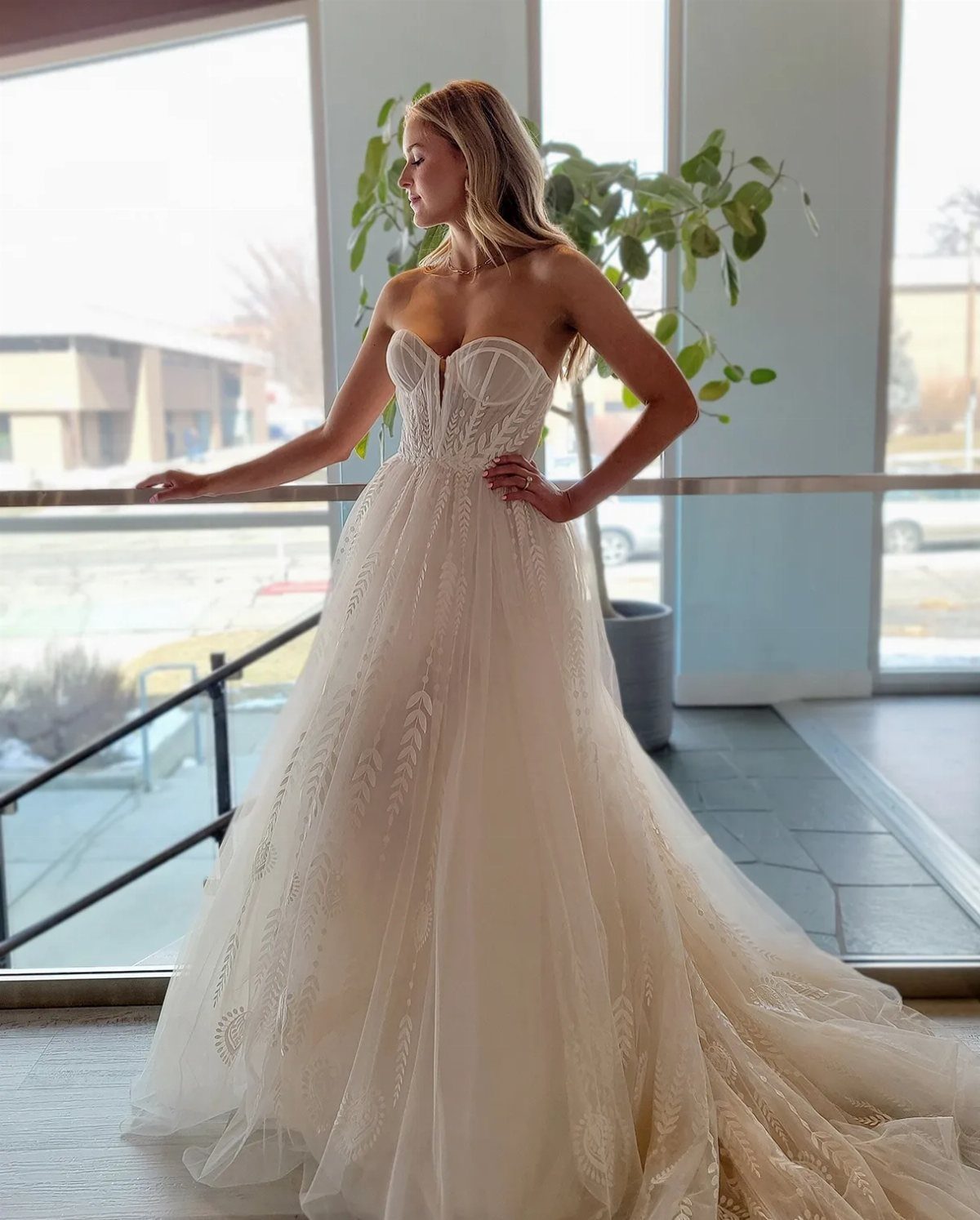 Woman wearing a strapless lace wedding dress with a voluminous tulle skirt standing by a window.