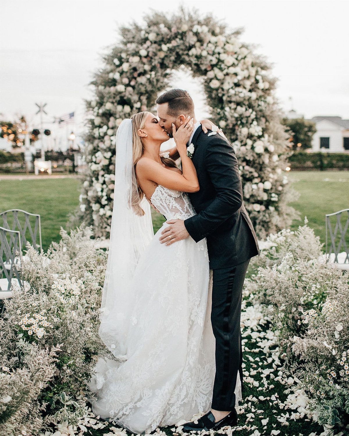 A bride and groom kissing centrally in front of a large white floral arch.