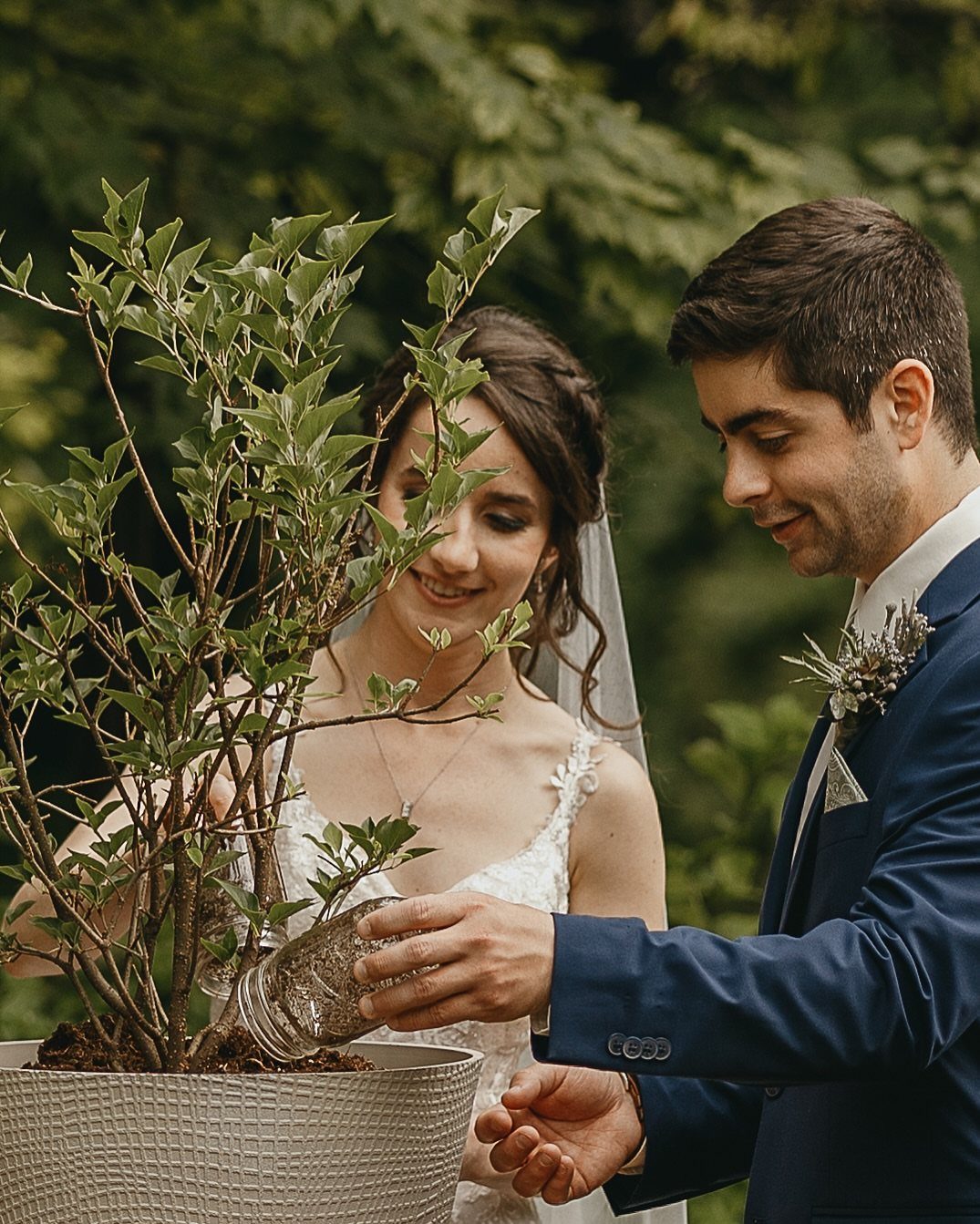 A bride and groom planting a small tree into a large grey pot outdoors.