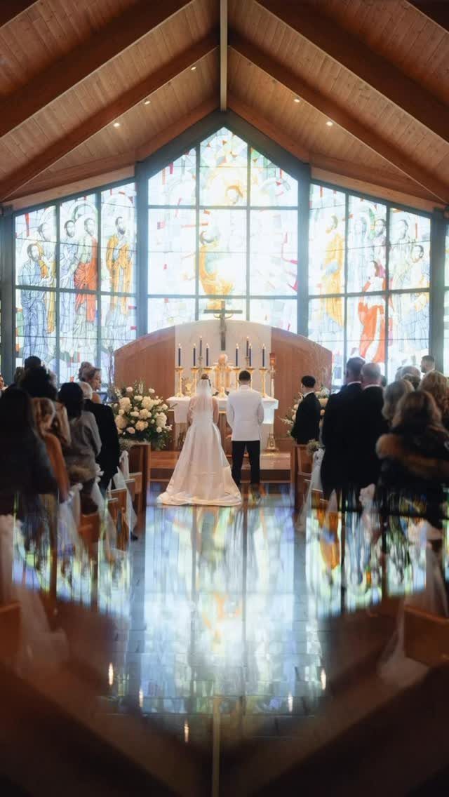 A bride and groom stand at a church altar before guests and stained glass windows.