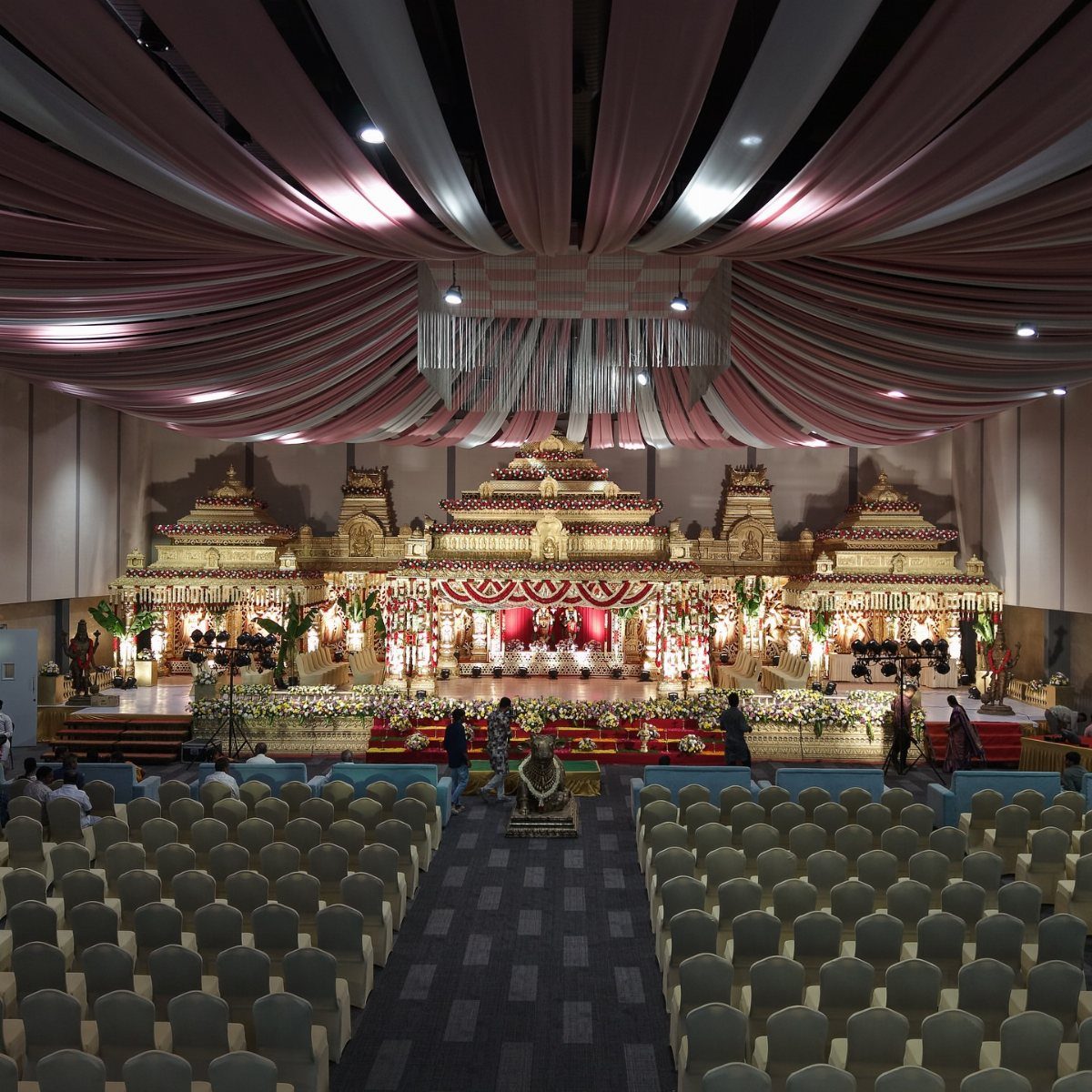 Ornate traditional Indian wedding stage with gold architecture, floral decorations, and rows of blue chairs.