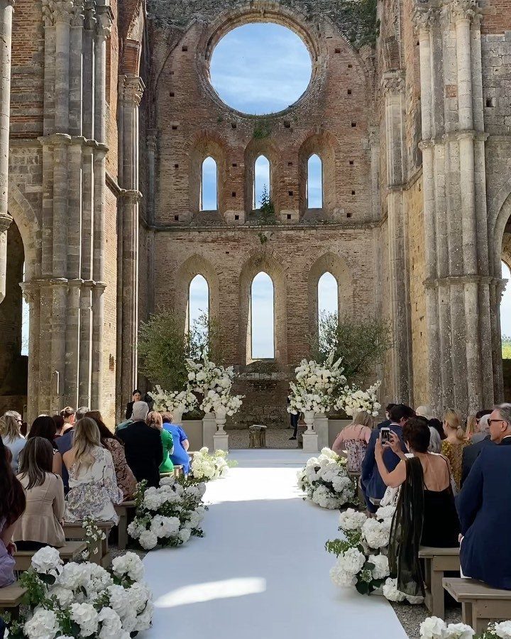 White floral arrangements line a wedding aisle leading toward ancient stone ruins under a blue sky.