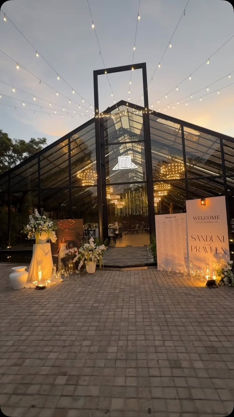 A glass pavilion wedding venue decorated with string lights and white floral arrangements at dusk.