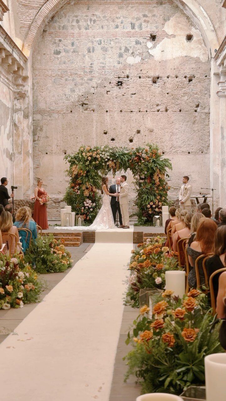 A bride and groom exchange vows under a floral arch in a rustic stone hall.