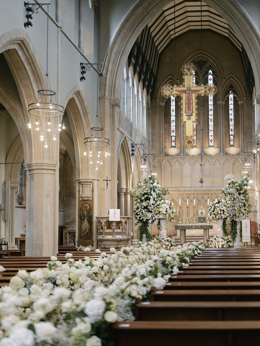 White floral arrangements line the wooden pews inside a stone church with hanging chandeliers.