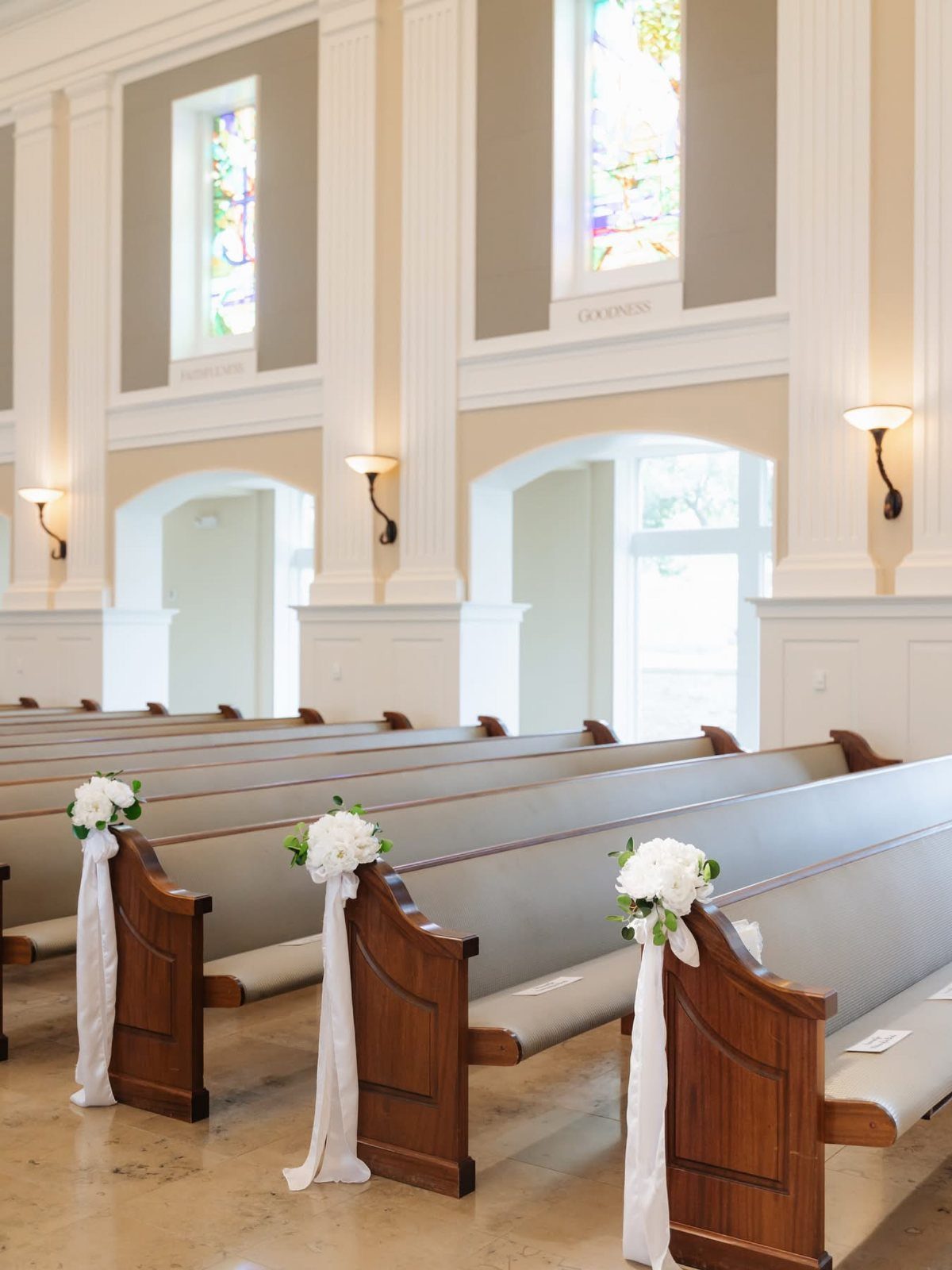 White floral arrangements with long flowing ribbons attached to the ends of wooden church pews.