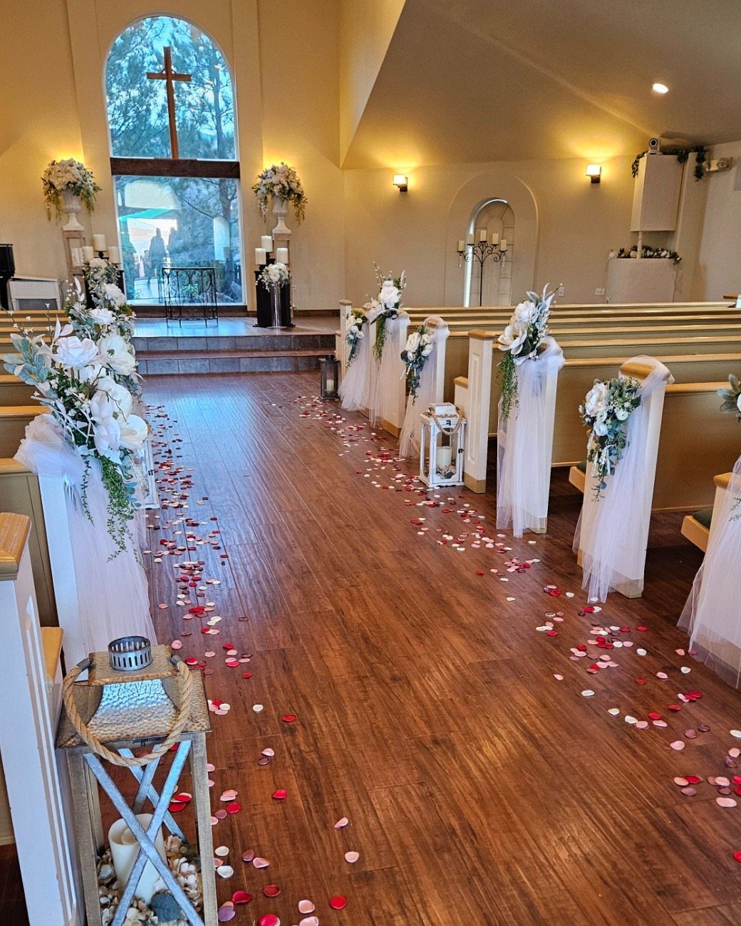 White tulle and floral arrangements decorate the wooden pews along a church aisle scattered with rose petals.