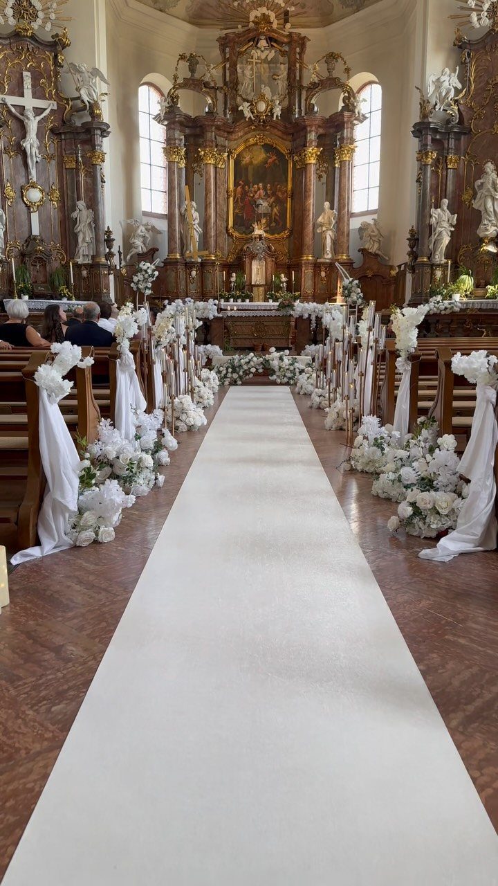 White aisle runner and floral decorations lining the pews in an ornate church interior.