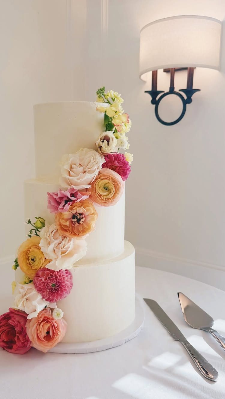 Wedding Decorations and Flowers: 8 Ideas for Ceremony to Reception 6 Three-tiered white wedding cake decorated with a cascading arrangement of colorful fresh flowers on a white table.