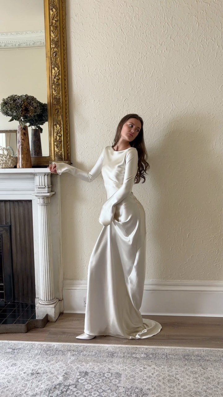 Wedding Dress Long Sleeve: 7 Styles From A-Line to Turtleneck 5 Woman in a long-sleeved white satin wedding dress standing by a white fireplace.