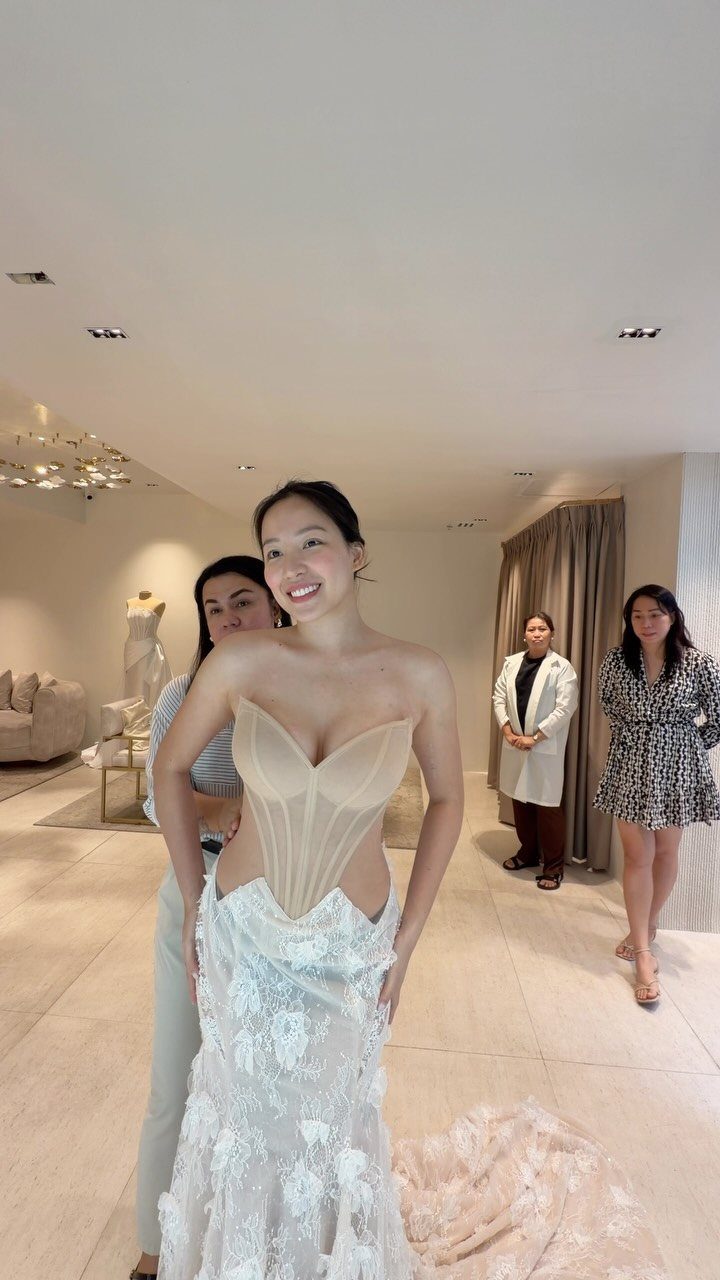 A woman smiles while trying on a white lace wedding dress with a nude corset bodice.