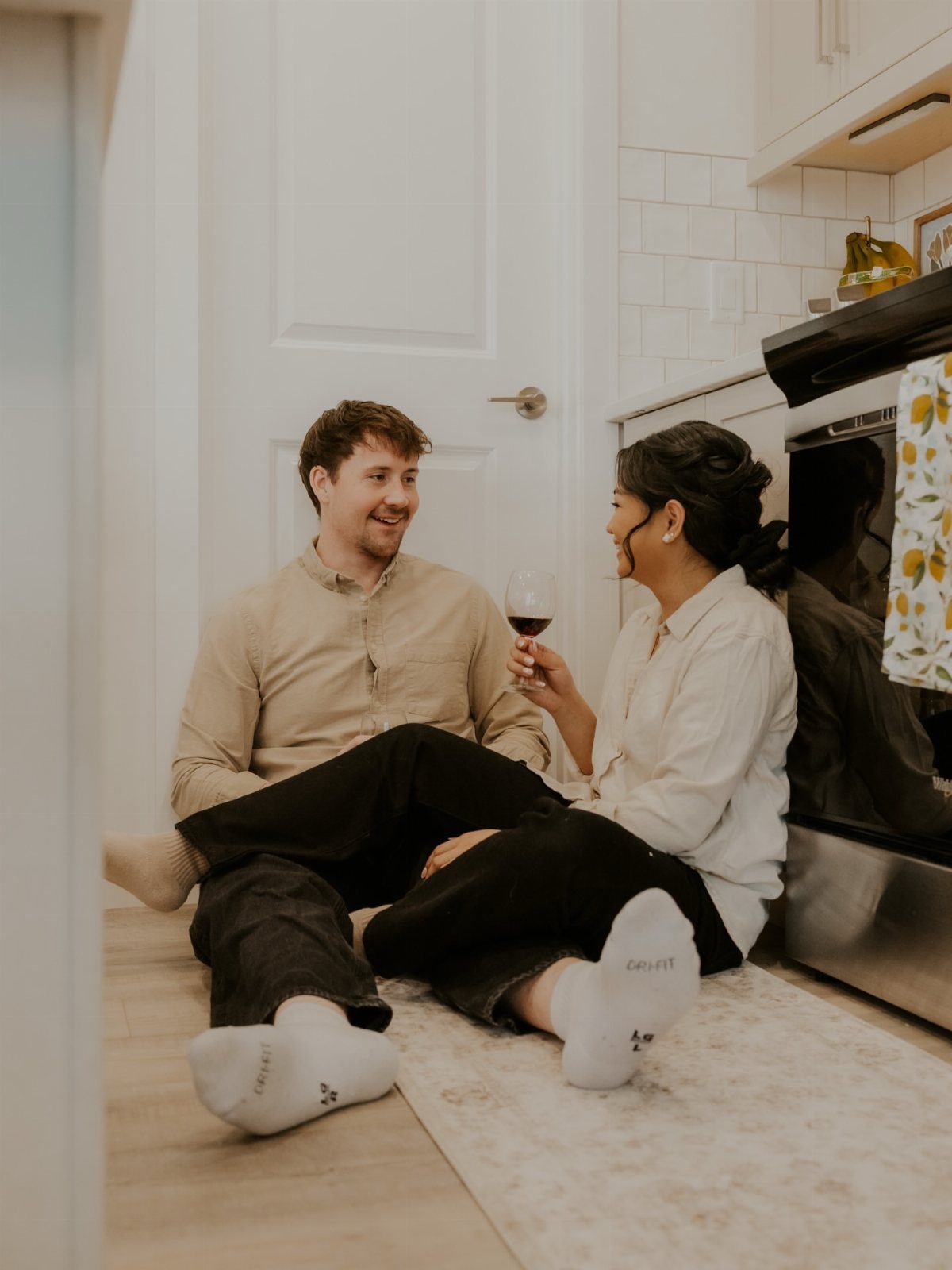 A man and woman sitting on a kitchen floor sharing a glass of red wine.