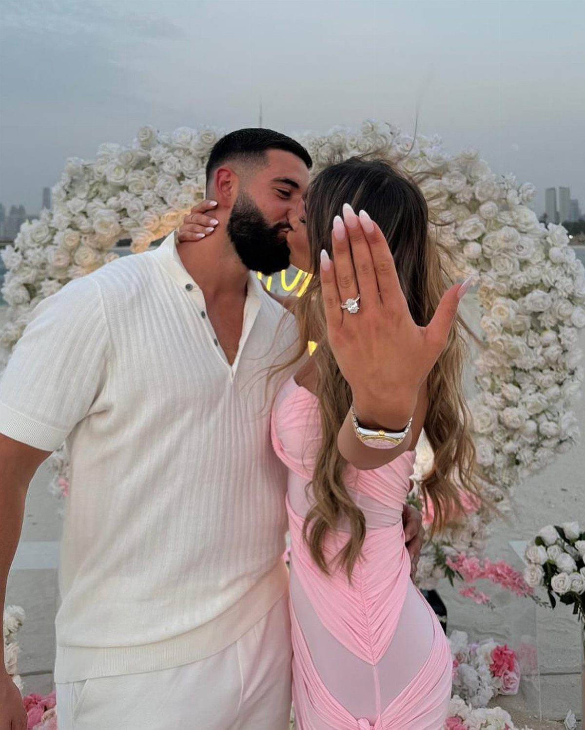 A woman displays her diamond engagement ring while kissing her partner in front of white flowers.