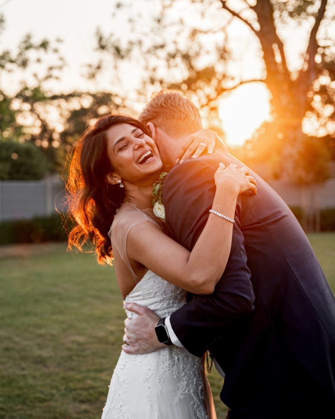 A laughing woman in a white dress hugging a man in a dark suit at sunset.