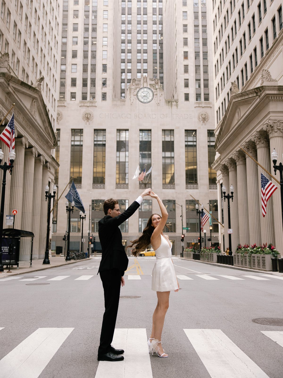 A couple dancing in a city street crosswalk surrounded by tall skyscrapers and flags.
