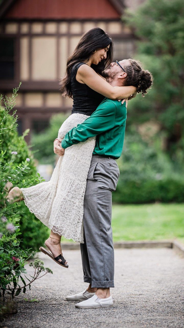 A man lifting a smiling woman in a white lace skirt during an outdoor engagement session.