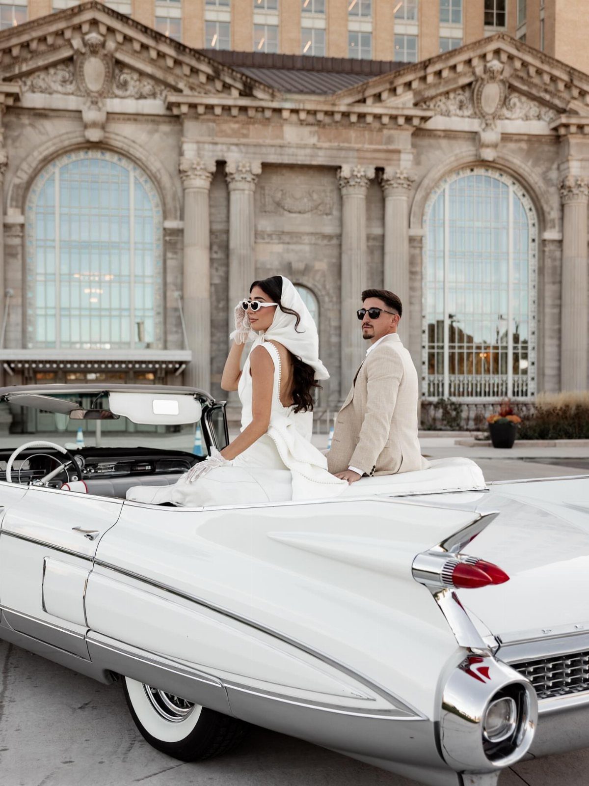 A couple in vintage attire posing in a white classic convertible car in front of a grand building.