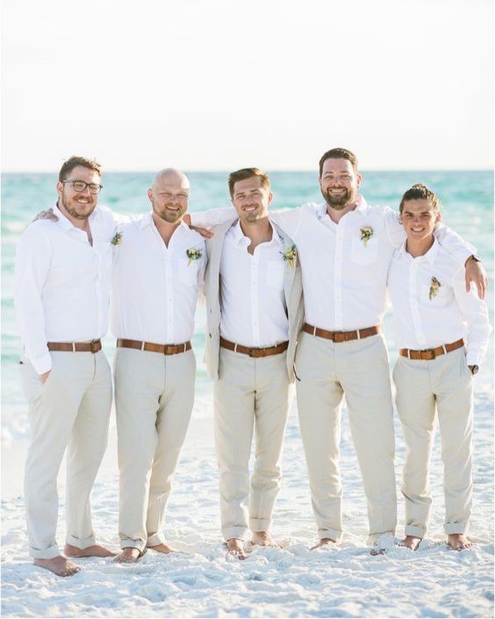 Five men in white shirts and beige trousers posing on a sunny beach.