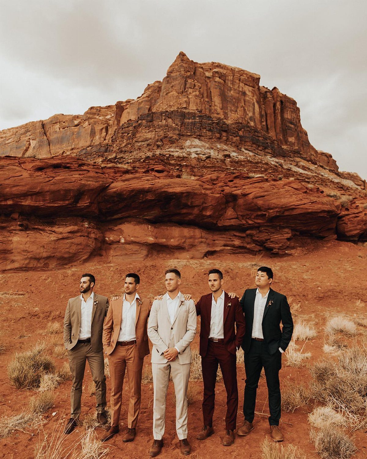 Five men standing in a desert landscape wearing various colored suits against red rock formations.