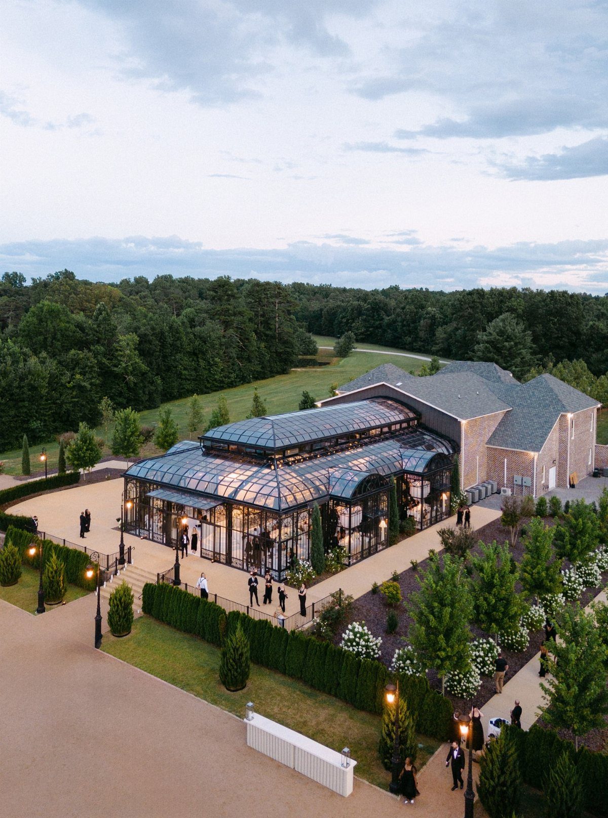 A large glass conservatory building surrounded by green lawns and manicured hedges at twilight.