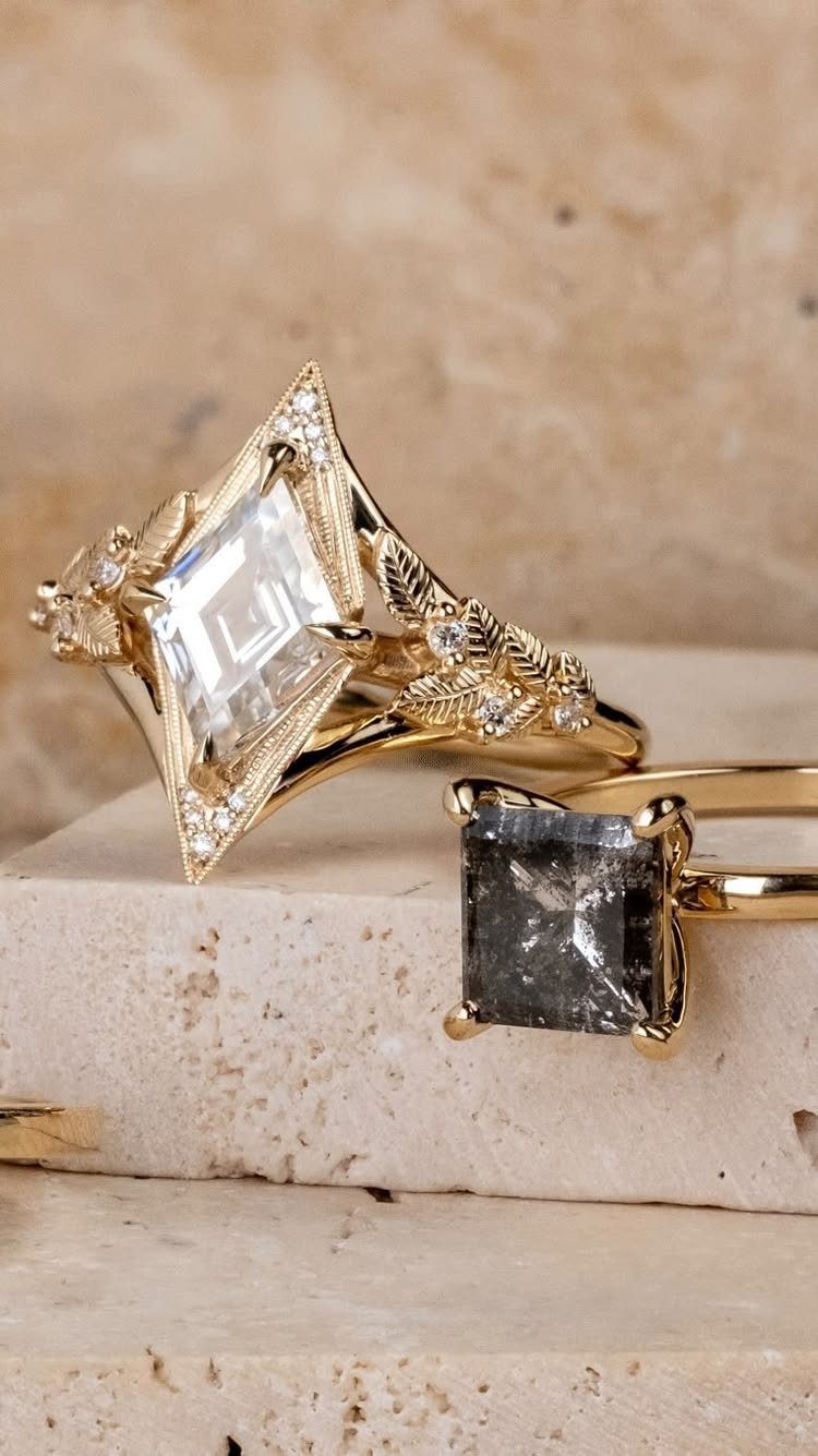 Two gold rings with leaf details and large gemstones resting on a beige stone surface.