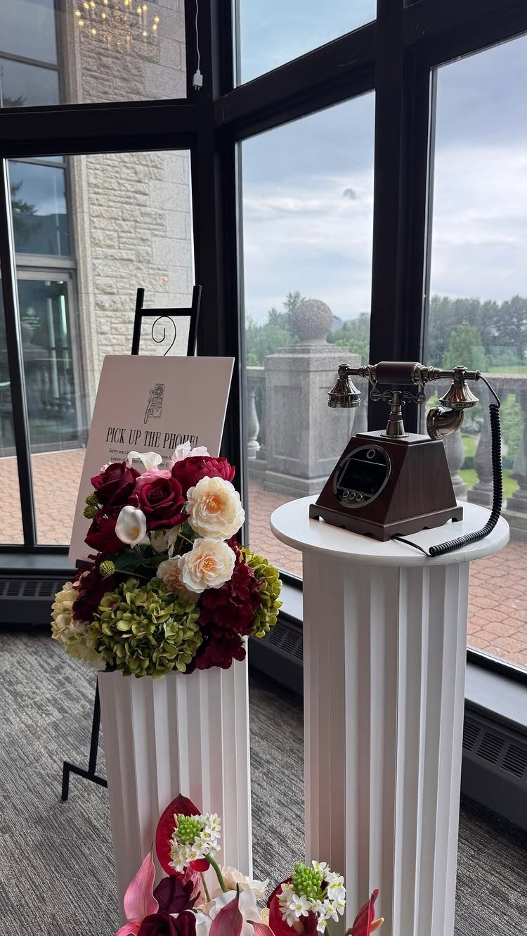 Vintage rotary telephone on a white pedestal next to floral arrangements and a sign near large windows.