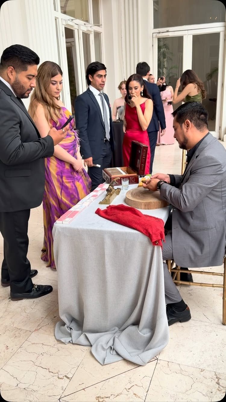 A professional cigar roller rolls cigars at a small table for wedding guests in formal attire.