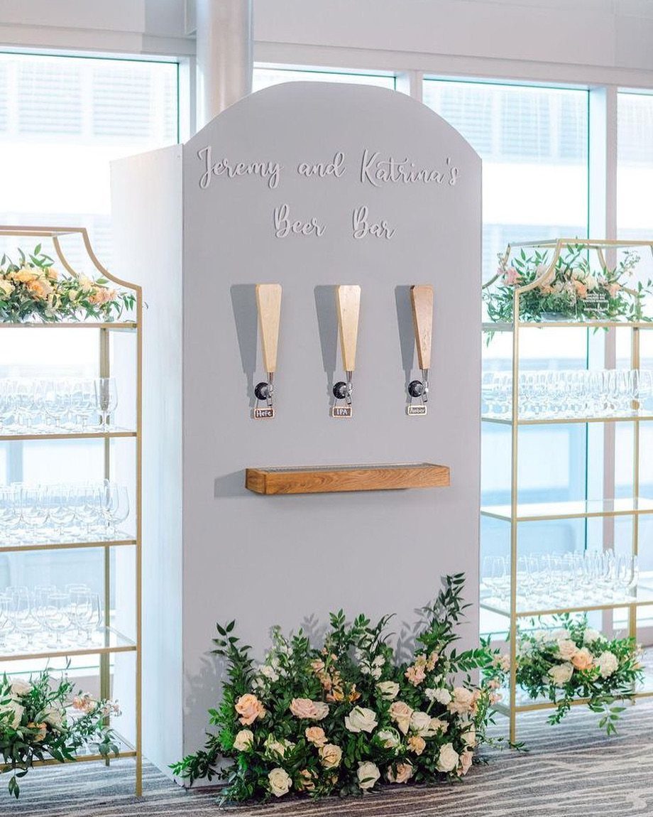 A custom grey beer tap station decorated with flowers and gold shelving units holding glassware.