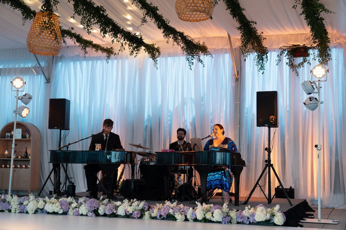 Two musicians playing grand pianos on a stage decorated with white flowers and greenery.