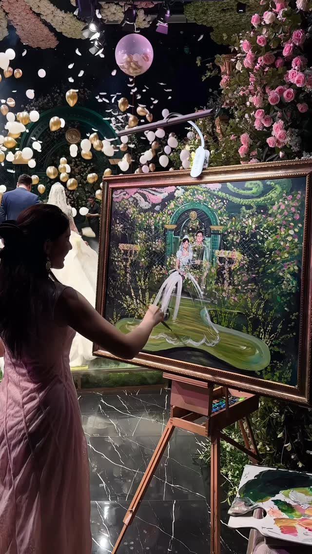 A female artist paints a wedding portrait of a bride and groom on a large canvas at an easel.