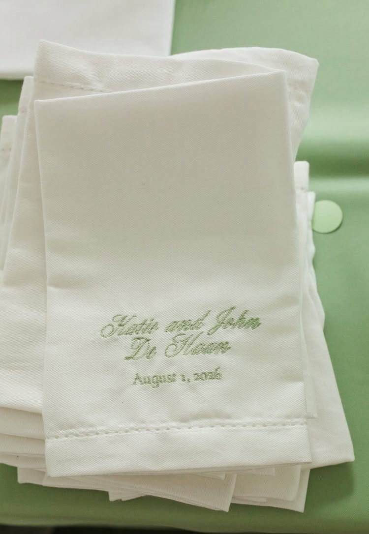 Stack of white embroidered napkins with names and a wedding date on a green background.