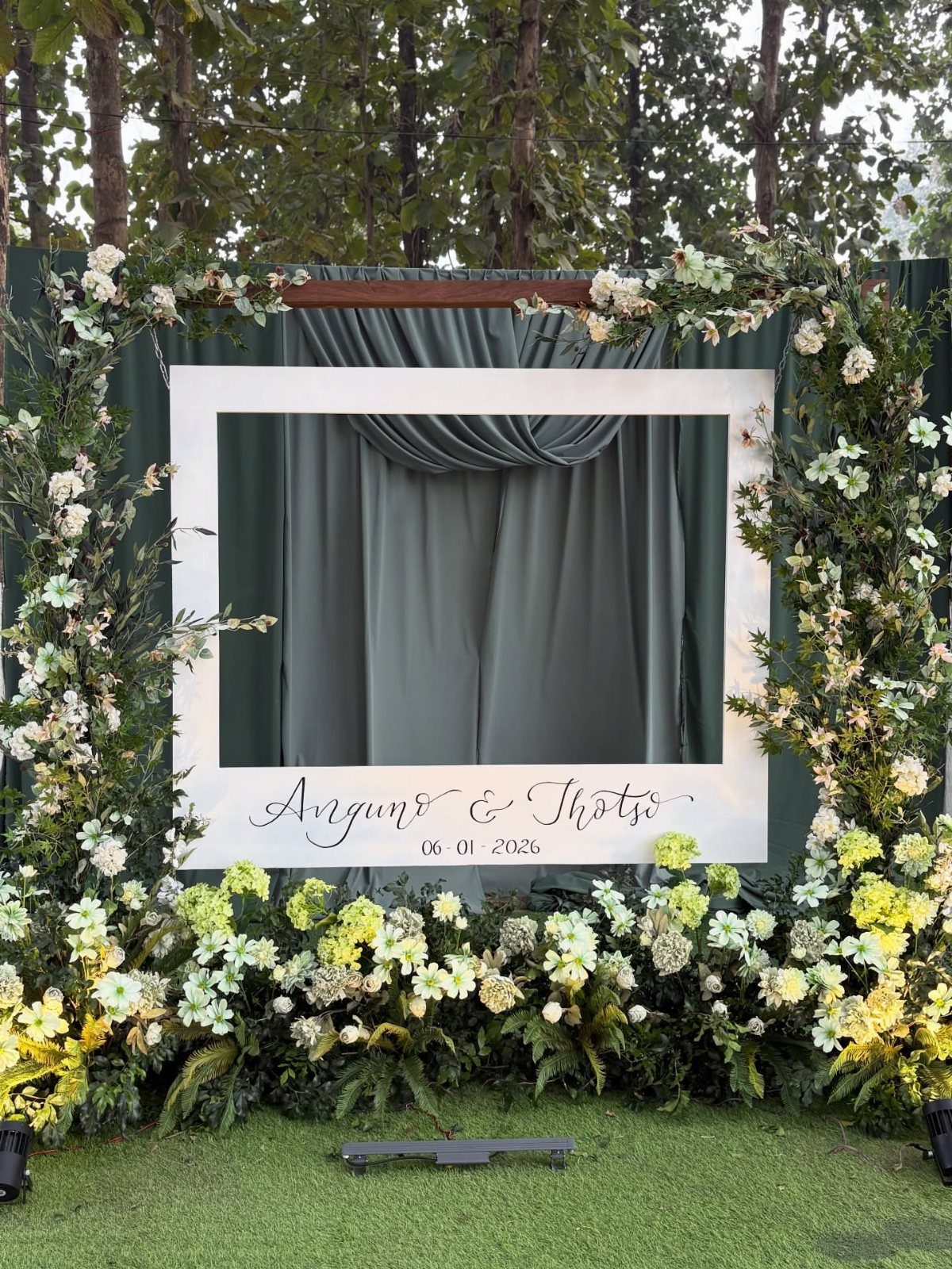 A large white floral frame with a grey fabric backdrop set on a green lawn.