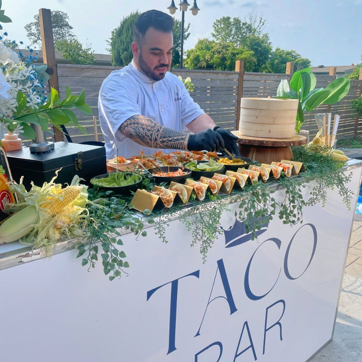 A chef prepares food at an outdoor taco bar station decorated with greenery and corn.