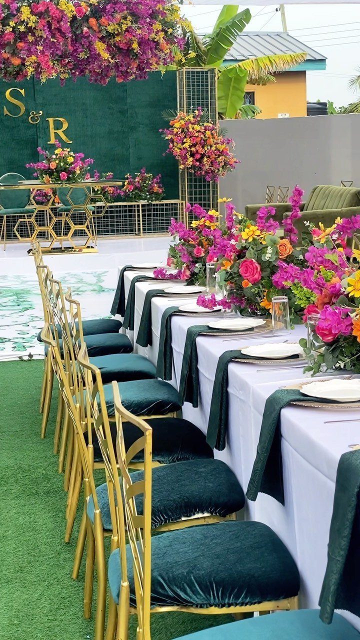 Long wedding table with gold chairs, emerald green velvet cushions, and bright pink floral arrangements.
