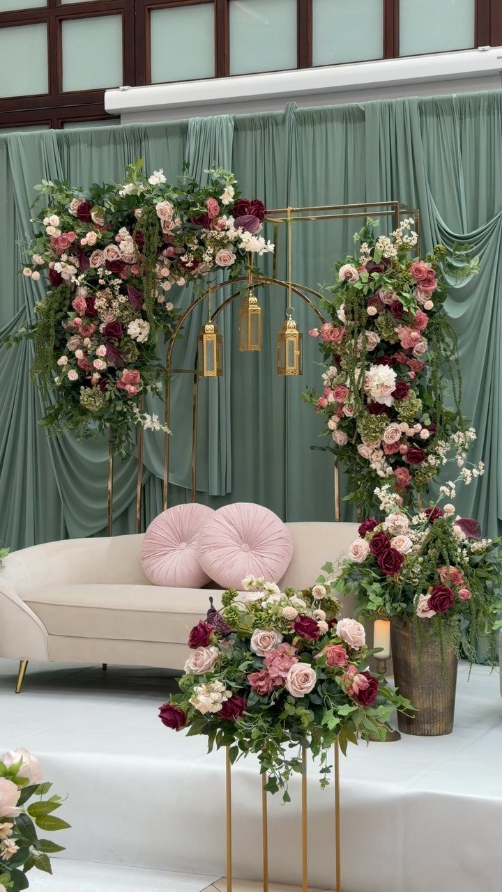 Floral wedding stage with pink and burgundy flowers, green foliage, and a cream sofa against green drapes.