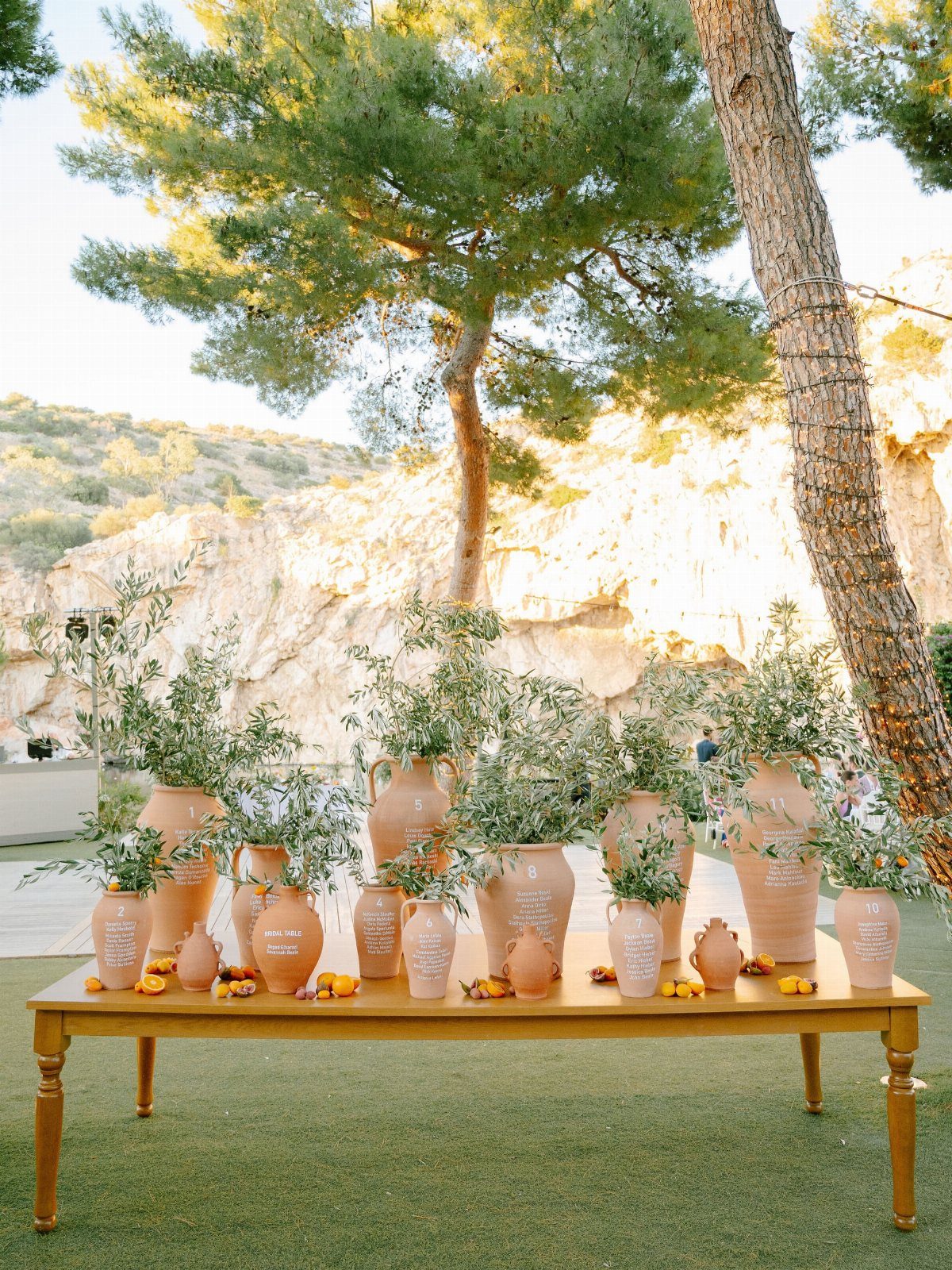 Terracotta vases filled with green leafy branches arranged on a long gold table outdoors.