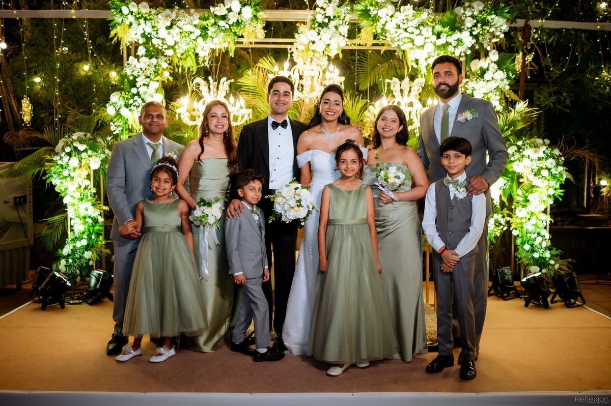 Wedding party posing in formal attire with bridesmaids wearing sage green dresses against a floral background.