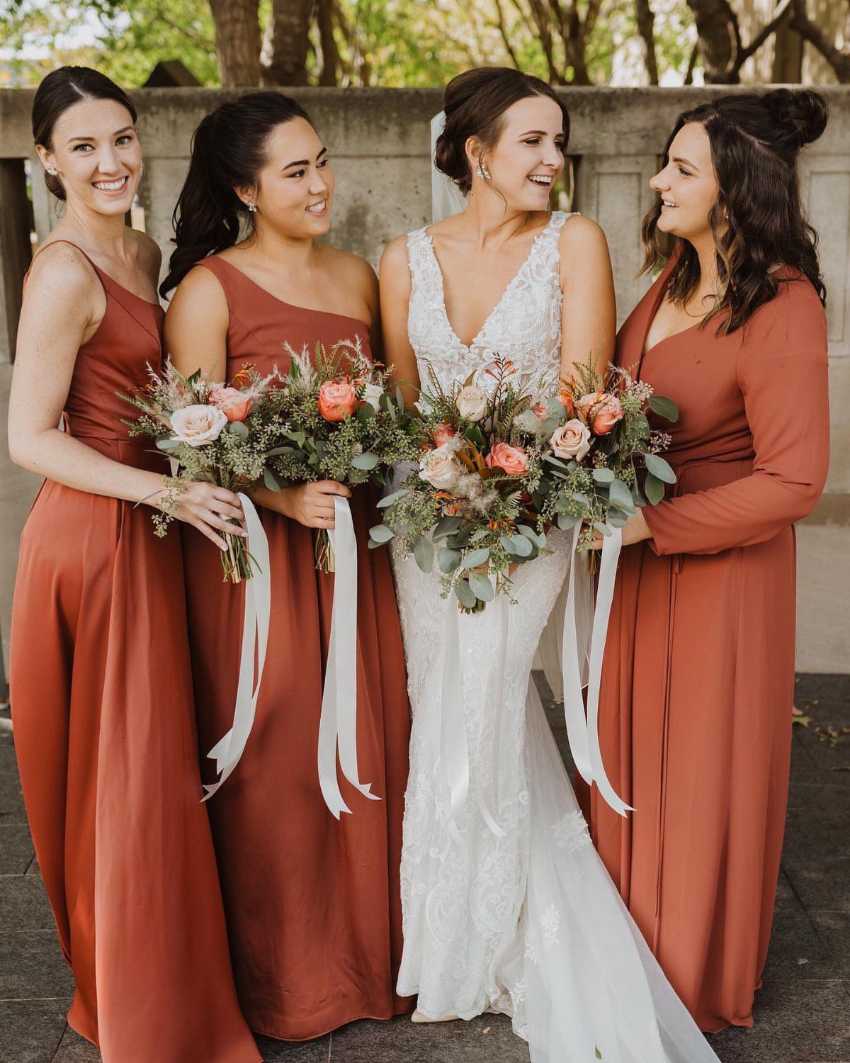 Three bridesmaids in cinnamon dresses standing with a bride in a white lace gown holding bouquets.