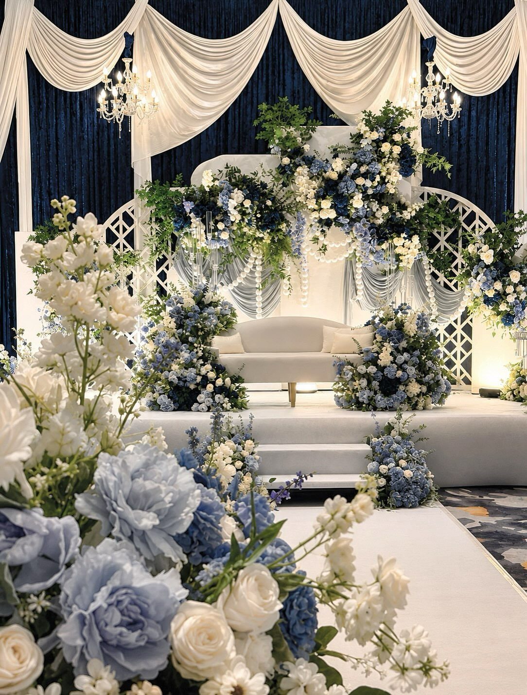 White and blue floral arrangements decorate a wedding stage with draped fabric and a white sofa.