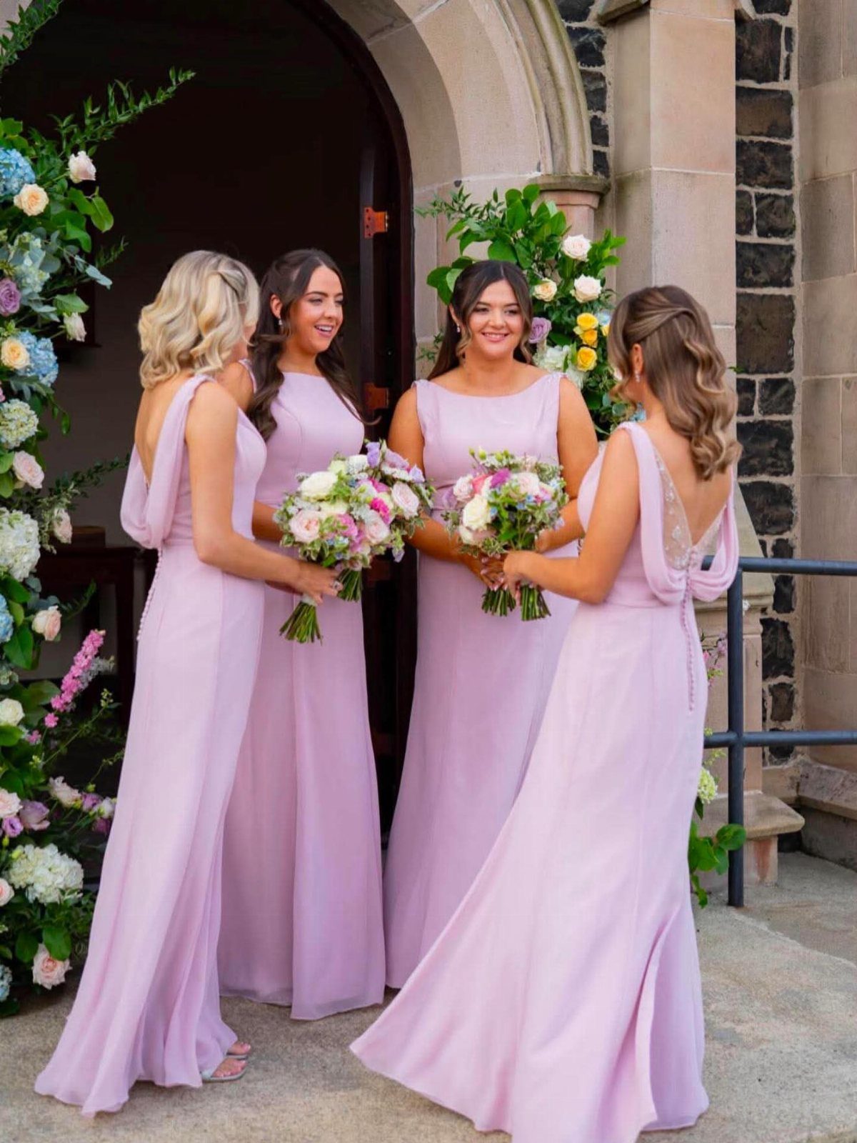 Four bridesmaids in lavender dresses holding bouquets in front of a stone archway.