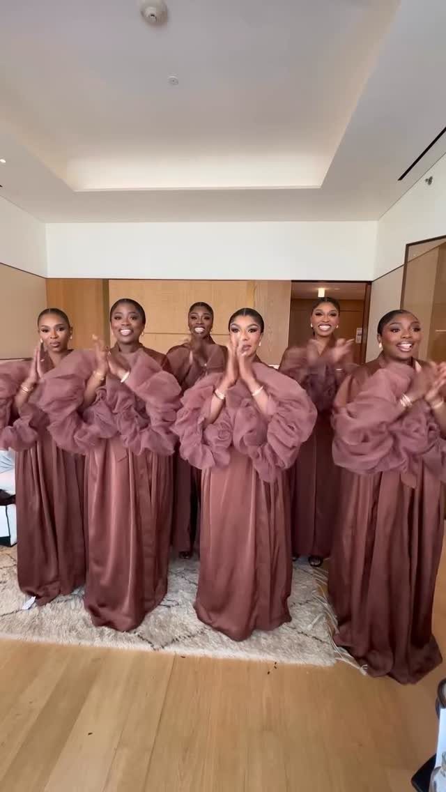 Six women posing in long, mauve satin dresses with large ruffled sleeves in a bright room.