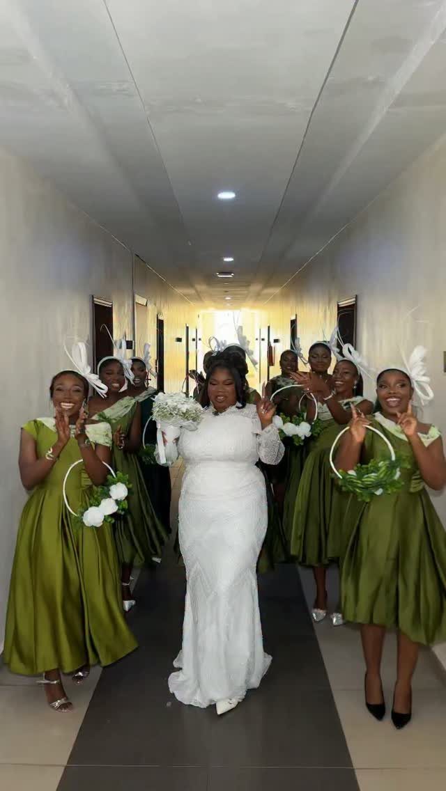 A bride in white walking through a hallway surrounded by bridesmaids in olive green dresses.
