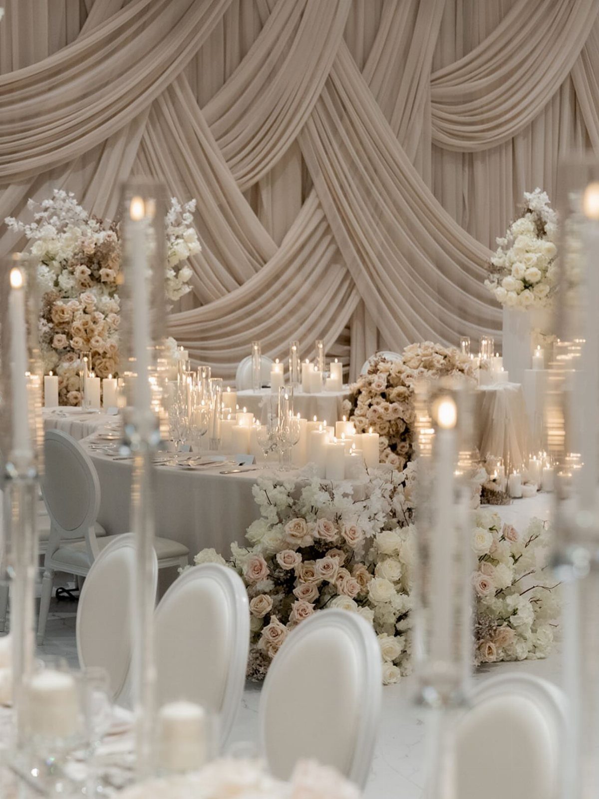 Wedding reception table decorated with white candles, cream roses, and draped beige fabric backdrop.