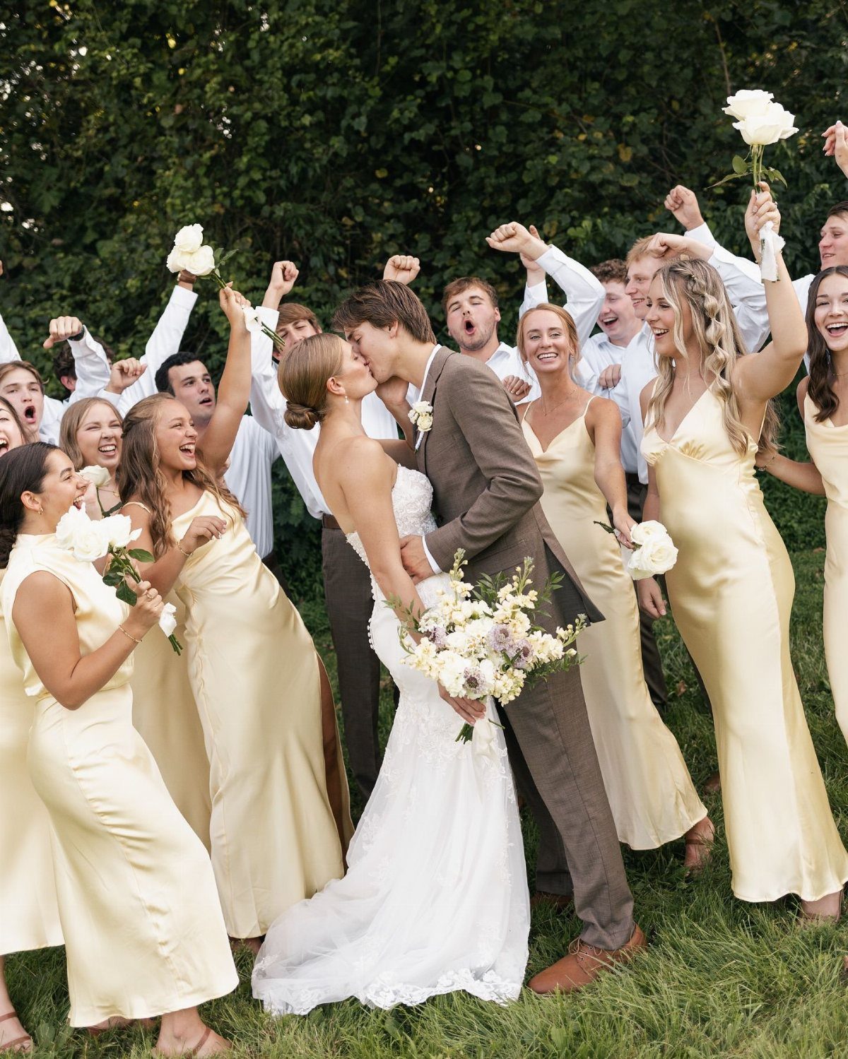 Bridesmaids in gold satin dresses cheer as a bride and groom kiss outdoors.
