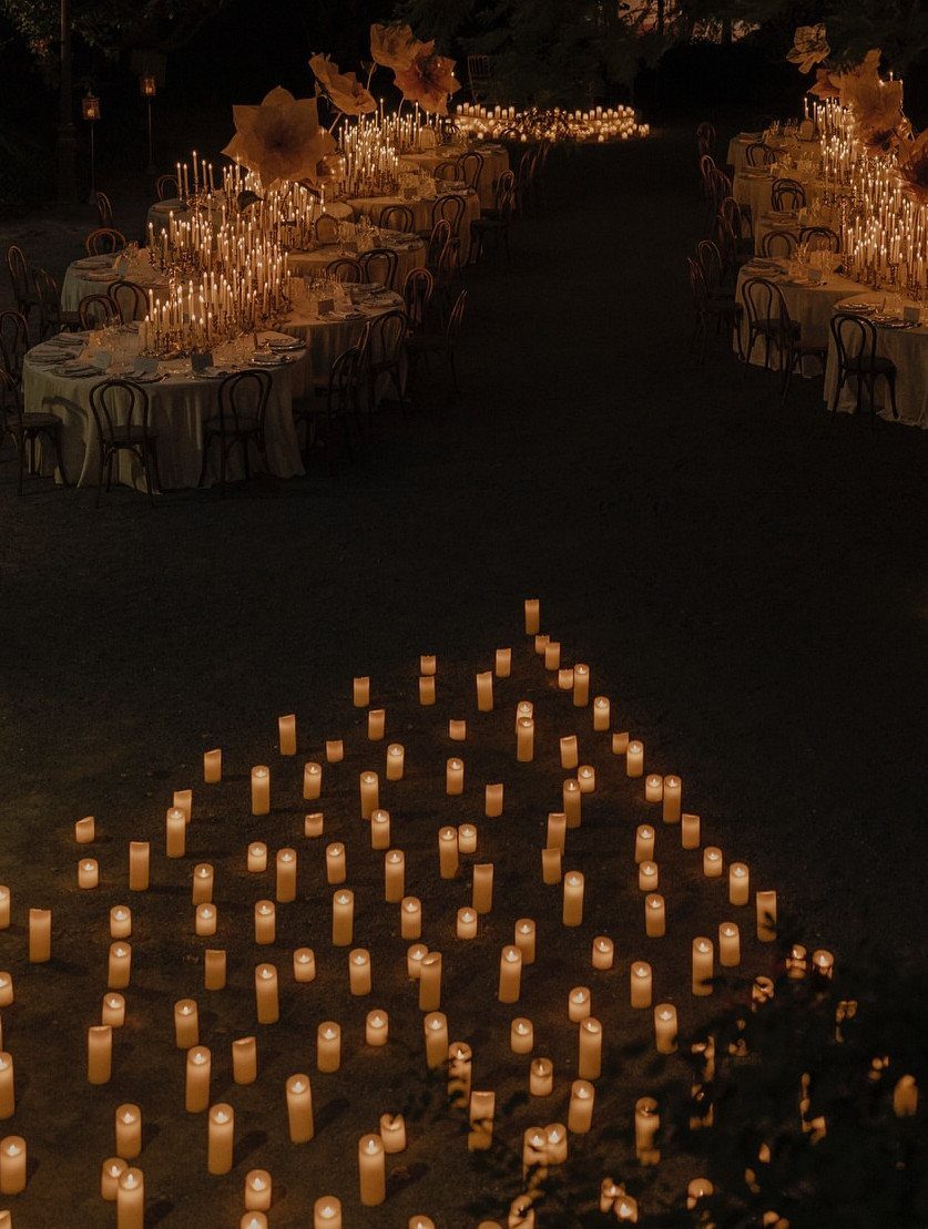 Small glowing candles scattered on the ground leading toward long, candlelit dining tables at night.